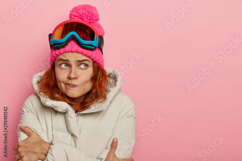 People, hobby and winter holidays concept. Unhappy young European woman trembles from cold, purses lips, spends much time during frosty weather, looks on right side, stands over rosy background