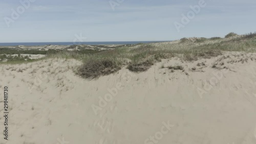 Drone shot flying forwards over some dunes, revealing more dunes and the ocean behind it. photo