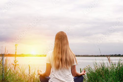 Beautiful girl with long hair is sitting on the shore. The view from the back. Sunset. Peace and tranquility. Yoga by the lake