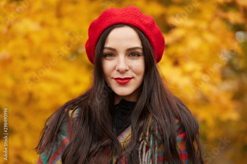 Facial portrait of beautiful Caucasian woman warmly clothed, posing in autumn outdoor, looking directly at camera with calm facial expression, lady dresses stylish red beret, spending time in open air