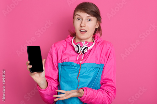 Cheerful pretty young Caucasian sportswoman holding blank screen smartphone and pointing on it isolated over pink background, having schocked facial expression, copy space for adertisment or promotion photo