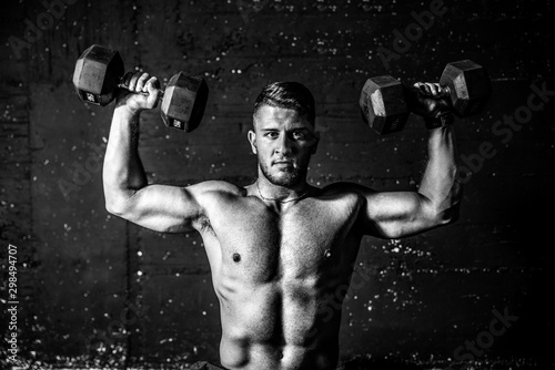 Young fit strong sweaty man shoulders workout training with two dumbbells in the gym dark image with shadows real people black and white