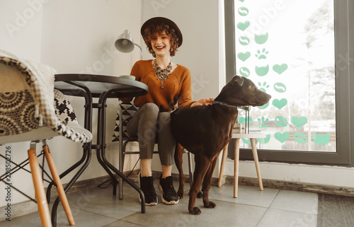 Portrait of happy lady in stylish clothes and hat sitting in chair in cozy cafe light, petting dog, looking at camera and smiling. Girl hipster with dog relaxing in atmospheric cafe.