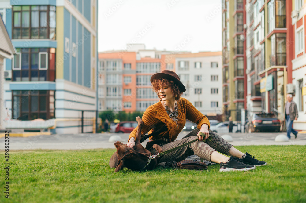 Fototapeta premium Stylish girl playing with a beautiful brown puppy on the lawn, smiling and stroking the dog. Beautiful lady is having fun on the grass with a dog in the yard on a background of a modern city block.