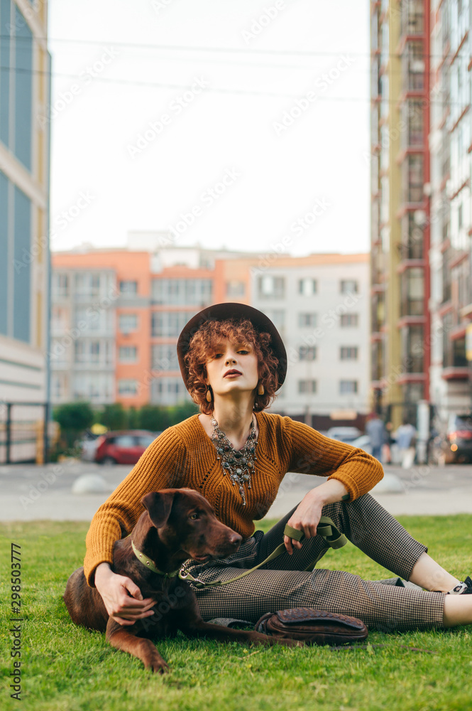 Beautiful girl with short hair and hat sits on the lawn, holds a dog on a leash and looks in camera, background of cityscape.Owner walks in with a cute brown dog sitting down to rest on the grass Beautiful girl with short hair and hat sits on the lawn, holds a dog on a leash and looks in camera, background of cityscape.Owner walks in with a cute brown dog sitting down to rest on the grass