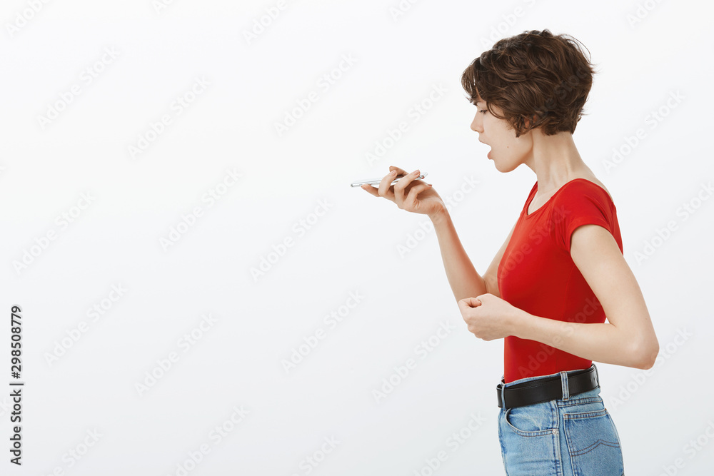 Angry intense serious girl stands profile hold smartphone near mouth record voice message clench fist outraged yelling talking speaker asking AI assistant start playing music white background Angry intense serious girl stands profile hold smartphone near mouth record voice message clench fist outraged yelling talking speaker asking AI assistant start playing music white background