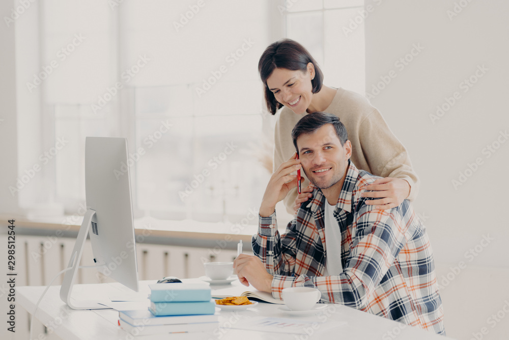 Obraz premium Caring woman helps her husband entrepreneur who has telephone conversation, tries to soleve working issues, writes information in notepad and works on computer. Family couple manage finances