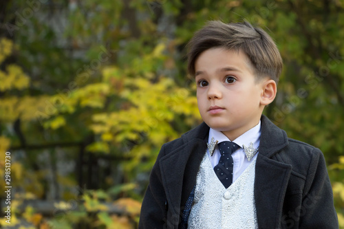 Portrait of a boy with surprise on his face on a background of autumn trees with copy space. A small man dressed in a business suit, a white shirt with a tie, coat. Emotional development in childhood