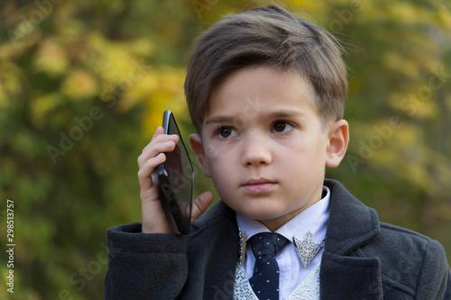Portrait of pensive little businessman talking by phone on autumn background. The development of modern children concept. Little adults. A small man dressed in a business suit, white shirt and tie