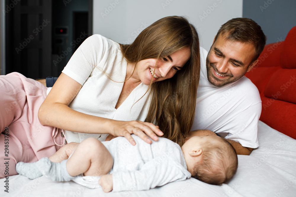 Young happy cheerful family of three lying in bed together. Parents play with their little baby Young happy cheerful family of three lying in bed together. Parents play with their little baby