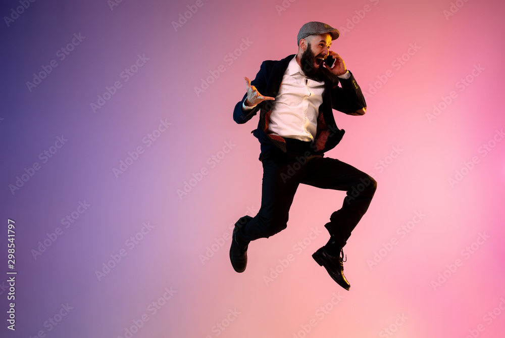 Full length portrait of happy jumping man wearing office clothes in neon light isolated on gradient background. Emotions, ad concept. Using smartphone, winning bet or sale, shopping, talking Full length portrait of happy jumping man wearing office clothes in neon light isolated on gradient background. Emotions, ad concept. Using smartphone, winning bet or sale, shopping, talking