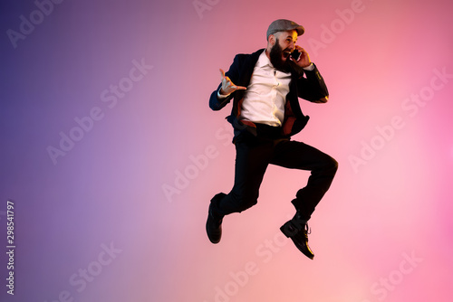 Full length portrait of happy jumping man wearing office clothes in neon light isolated on gradient background. Emotions, ad concept. Using smartphone, winning bet or sale, shopping, talking