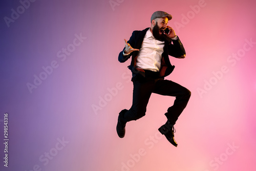 Full length portrait of happy jumping man wearing office clothes in neon light isolated on gradient background. Emotions, ad. Using smartphone, winning bet or sale, shopping, hurrying up, office work.