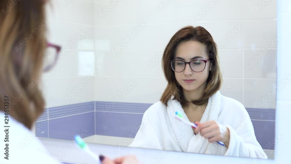 A young woman in glasses brushes her teeth. A woman in a white coat began to brush her teeth. View through the mirror A young woman in glasses brushes her teeth. A woman in a white coat began to brush her teeth. View through the mirror