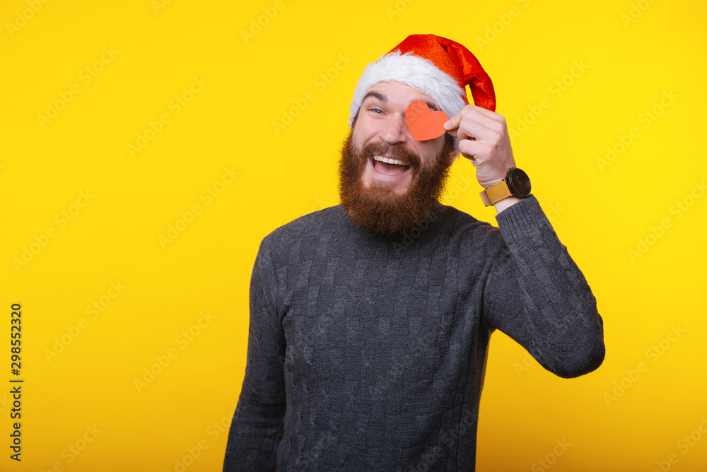 Image of funny bearded man wearing santa claus hat and put red heart shape over eye Image of funny bearded man wearing santa claus hat and put red heart shape over eye