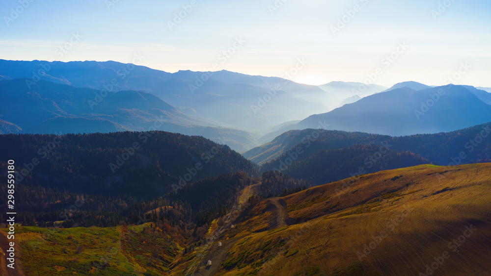 Beautiful aerial view to the evening mountains in Sochi Roza Hutor. Picturesque motion of autumn valley with the sun behind the peaks and hazy sunshine.