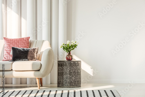 White flowers on glass vase on stylish table next to white sofa with pastel pink and black velvet pillows, real photo with copy space on empty white wall