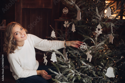 Full length of dark-haired woman in warm hat and socks in white sweater sitting on wooden porch with mug of hot drink or cacao in beautifully decorated room with Christmas tree and garland. It is
