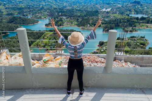 Vista trasera de mujer turista con sombrero levantando las manos al borde de la piedra del Peñol mientras contempla la naturaleza en Guatape Colombia photo