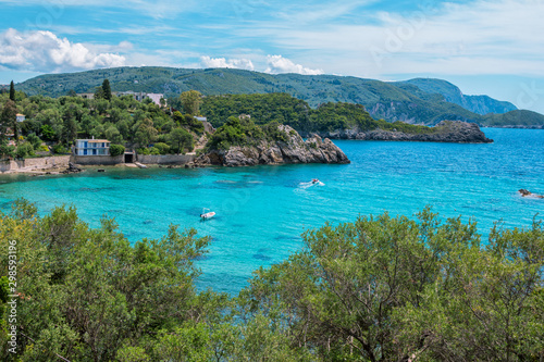 Landscape with turquoise calm sea water, mountain with rocky hillside covered with green trees and bushes and caves, cruise touristic boats and clouds on the sky. Corfu Island, Greece. 