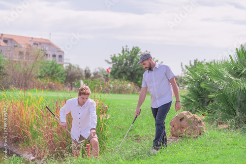 Two elegant men look for a golf ball on the shore of the lake on a golf course on a sunny day. Concept of relaxation and leisure.