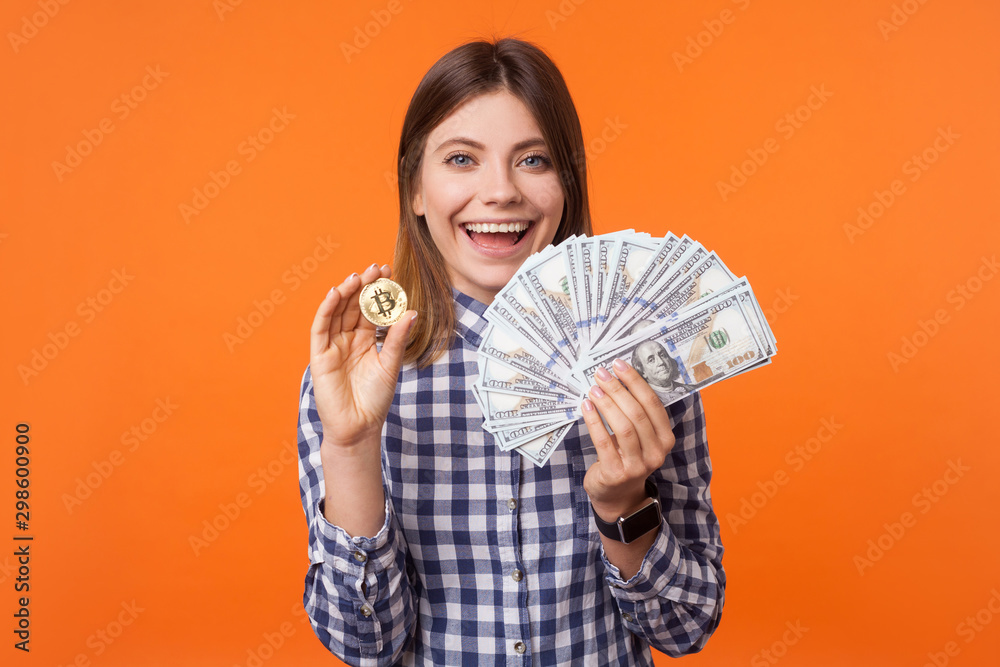 Portrait of excited brunette woman with charming smile wearing checkered shirt standing holding physical bitcoin and dollars in hands, cryptocurrency. indoor studio shot isolated on orange background Portrait of excited brunette woman with charming smile wearing checkered shirt standing holding physical bitcoin and dollars in hands, cryptocurrency. indoor studio shot isolated on orange background
