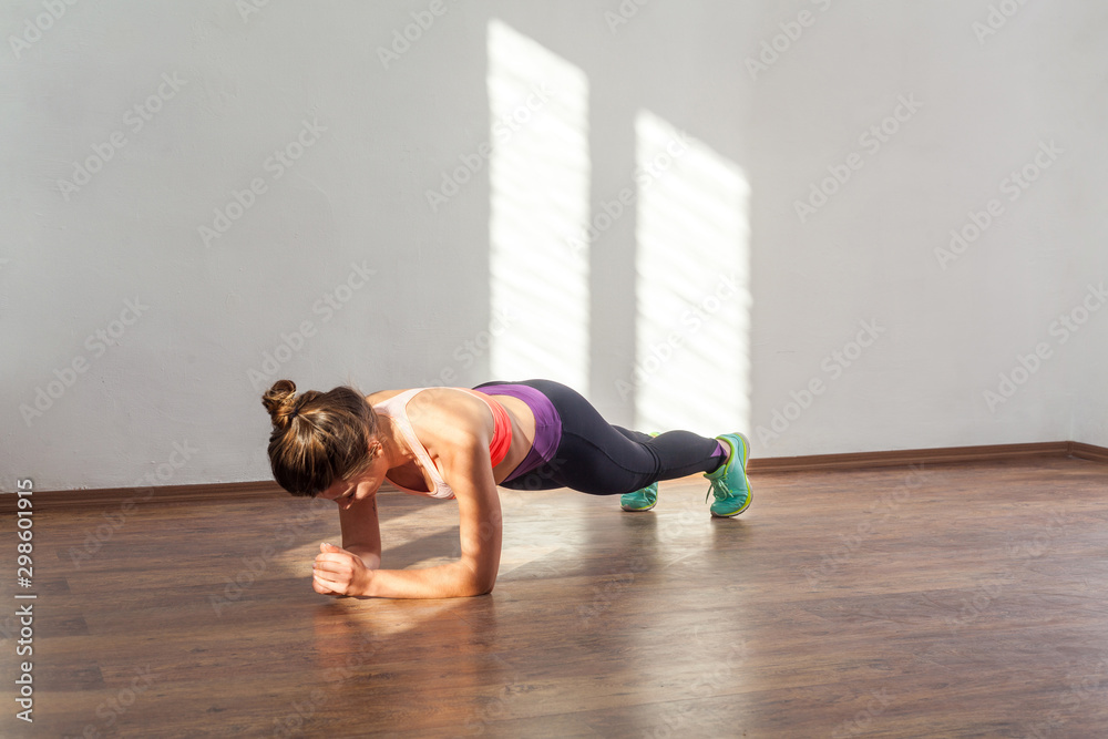 Athletic young woman with bun hairstyle and in sportswear practicing, doing plank exercise on elbows at home or fitness gym, looking down. indoor studio shot illuminated by sunlight from window