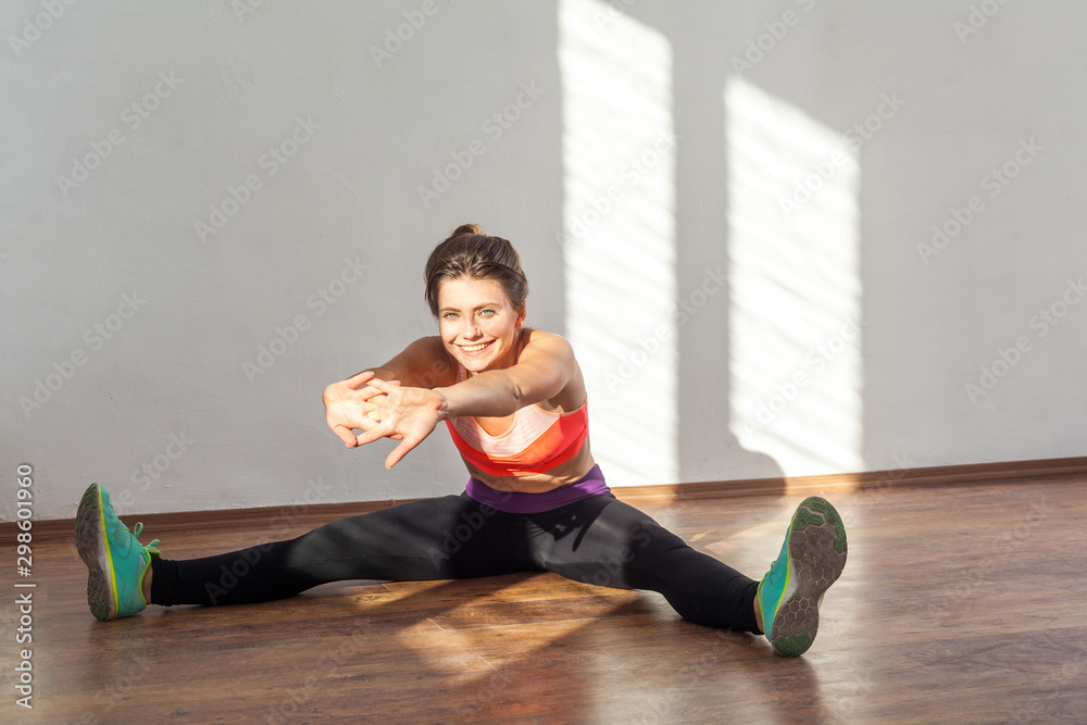 Beginner stretching, warm-up. Positive woman with bun hairstyle and in sportswear doing easy stretches and smiling at camera, practicing at home. indoor studio shot illuminated by sunlight from window