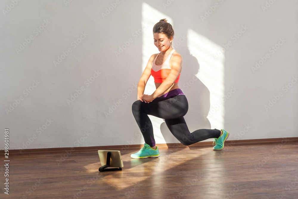 Diligent positive fit woman with bun hairstyle and in tight sportswear doing lunge or split squats while watching training video on tablet. indoor studio shot illuminated by sunlight from window Diligent positive fit woman with bun hairstyle and in tight sportswear doing lunge or split squats while watching training video on tablet. indoor studio shot illuminated by sunlight from window