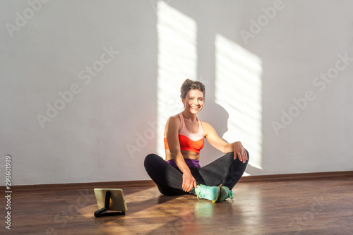 Charming fit woman with bun hairstyle and in tight sportswear sitting on floor, tired after workouts, tablet on floor showing training videos. indoor studio shot illuminated by sunlight from window