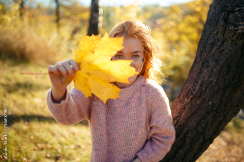 Portraits of a charming red-haired girl with a cute face. Girl posing in autumn park in a sweater and a coral-colored skirt. In the hands of a girl a yellow leaf