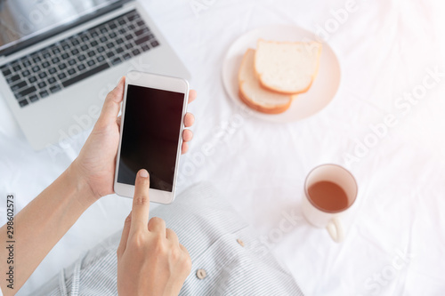 Hand of working woman holding and touching the black smartphone screen. Asian woman sitting on a white cloth while having bread, coffee and laptop.