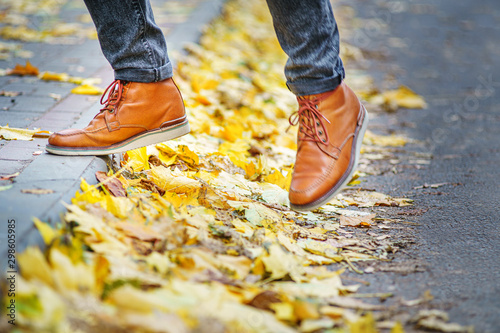 legs of a man in brown boots walking along the sidewalk strewn with fallen leaves. The concept of turnover of the seasons of the year. Weather background