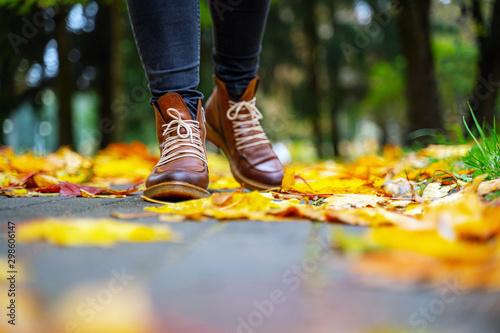 legs of a woman in black pants and brown boots walking in a park along the sidewalk strewn with fallen leaves. The concept of turnover seasons. Weather background