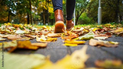 Back view on the feet of a woman in black pants and brown boots walking in a park along the sidewalk strewn with fallen leaves. The concept of turnover seasons. Weather background