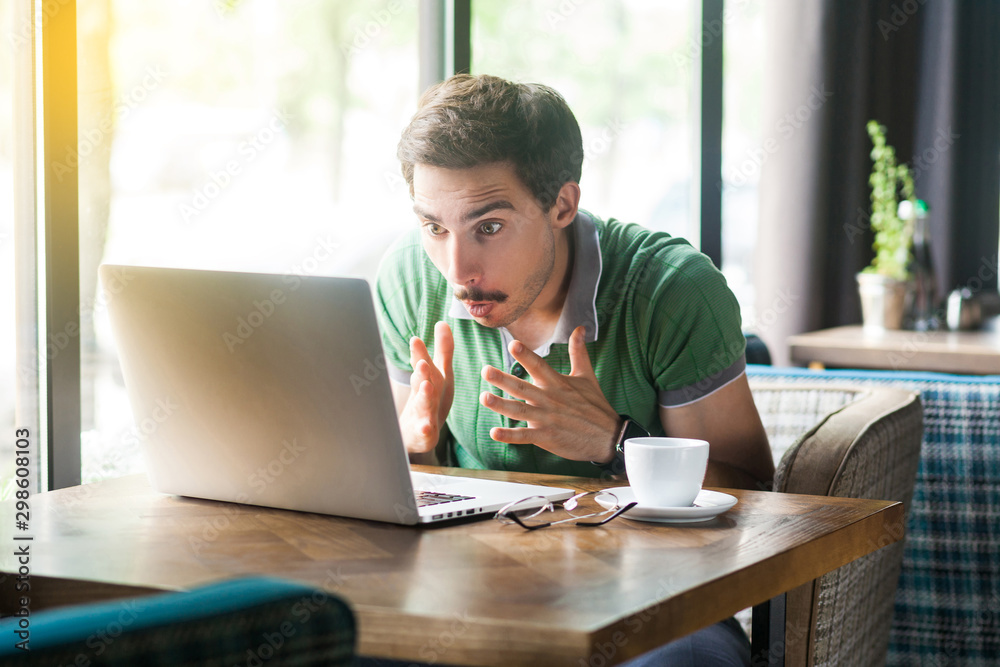 Oh my God! Young shocked businessman in green t-shirt sitting, looking at some unbelievable news at laptop screen and surprised. business and freelancing concept. indoor shot near window at daytime.