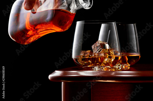 Two glasses of whiskey and ice on a wooden table in a low key. On top of the glass a man's hand with a bottle of whiskey.