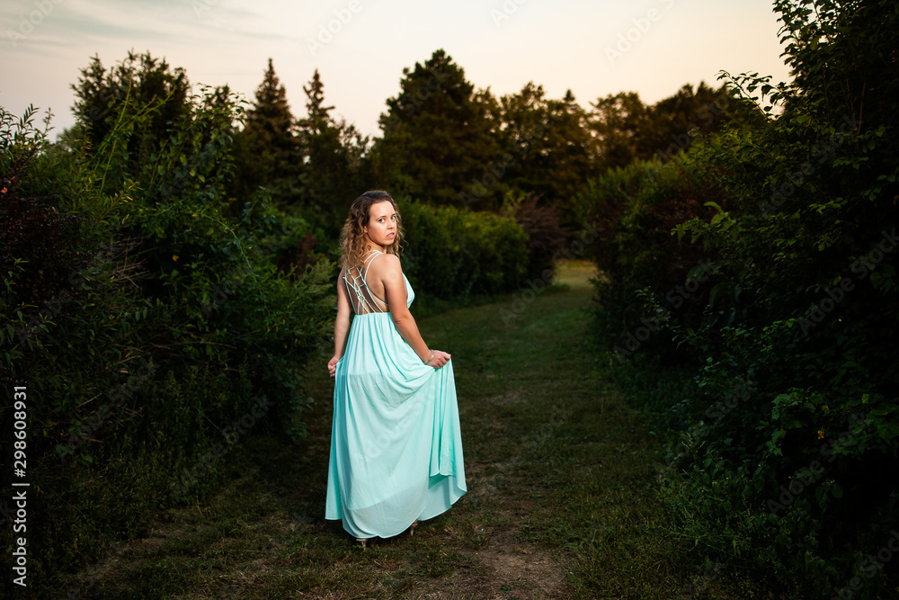 Beautiful curly hair young woman wearing elegant dress standing in the middle of forest with rays of sunlight during sunset Beautiful curly hair young woman wearing elegant dress standing in the middle of forest with rays of sunlight during sunset