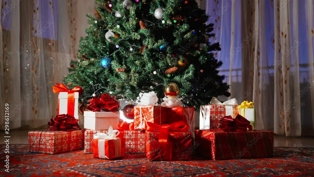 Flashing colorful christmas garland on the tree, round plastic balls and toys hanging on branches. Winter ornaments and decorations at home on window background. Xmas gift boxes lying under the tree