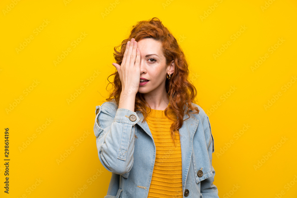 Redhead woman over isolated yellow background covering a eye by hand Redhead woman over isolated yellow background covering a eye by hand