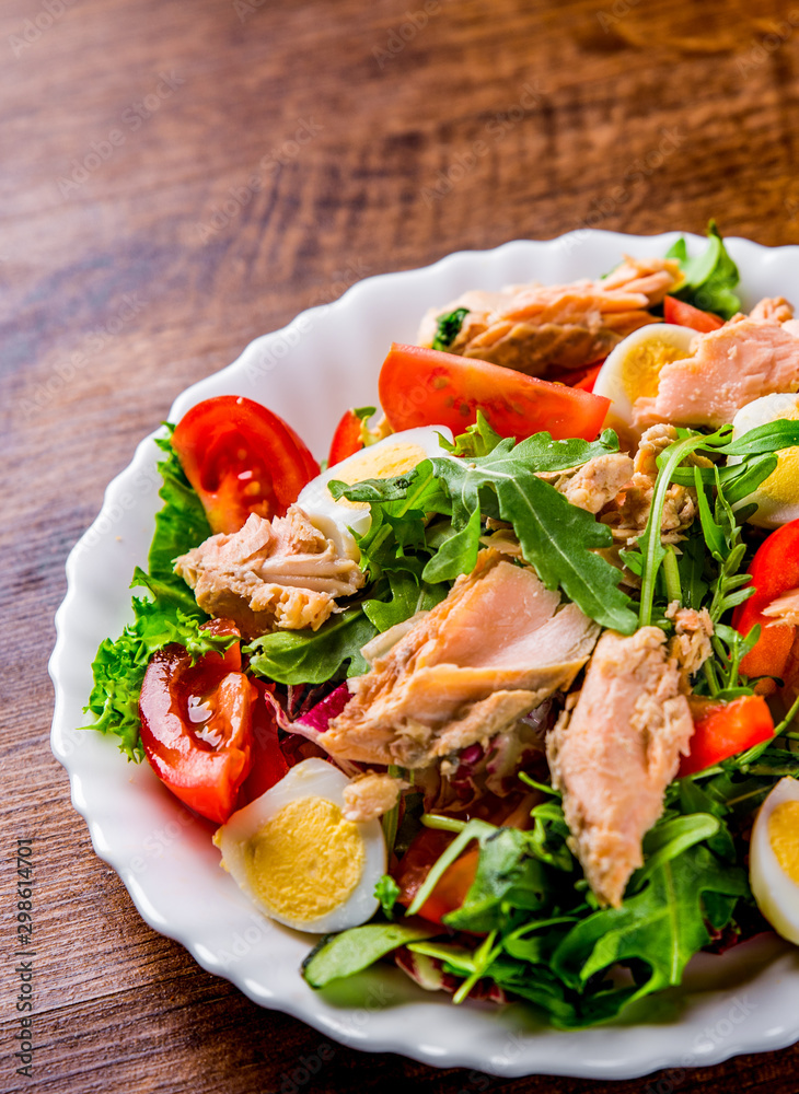 Fresh salad with fish, arugula, eggs,red pepper, lettuce, fresh sald leaves and tomato on a white plate on wooden table background
