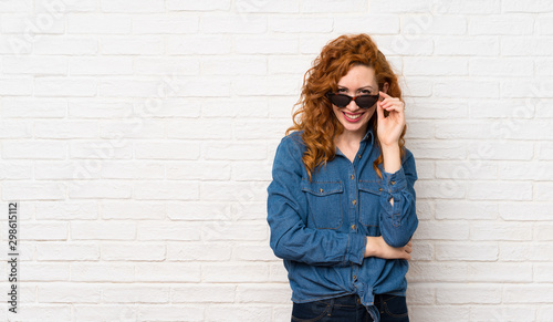 Redhead woman over white brick wall with glasses and happy