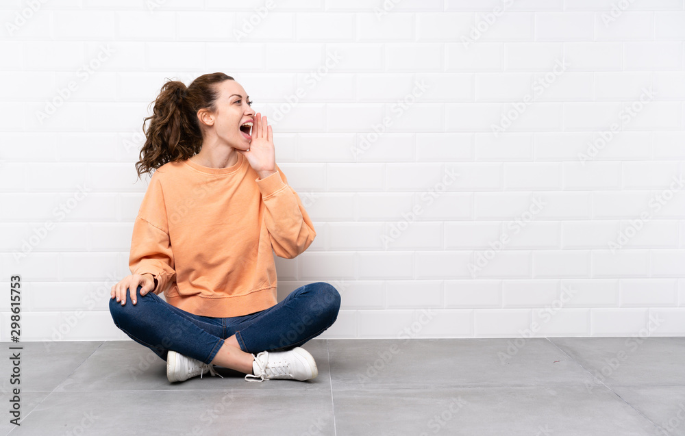 Young woman with curly hair sitting on the floor shouting with mouth wide open