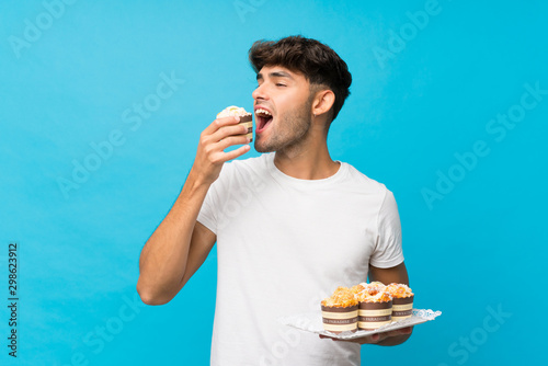 Young handsome man over isolated blue background holding mini cakes and eating it