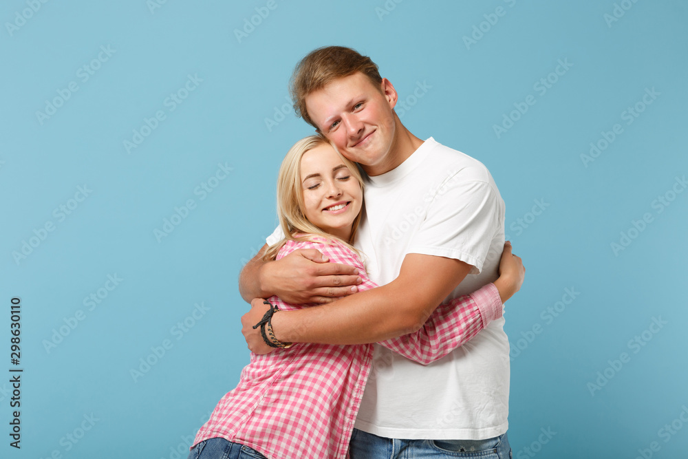 Young fun couple two friends guy girl in white pink empty blank design t-shirts posing isolated on pastel blue background studio portrait. People lifestyle concept. Mock up copy space. Hugging love