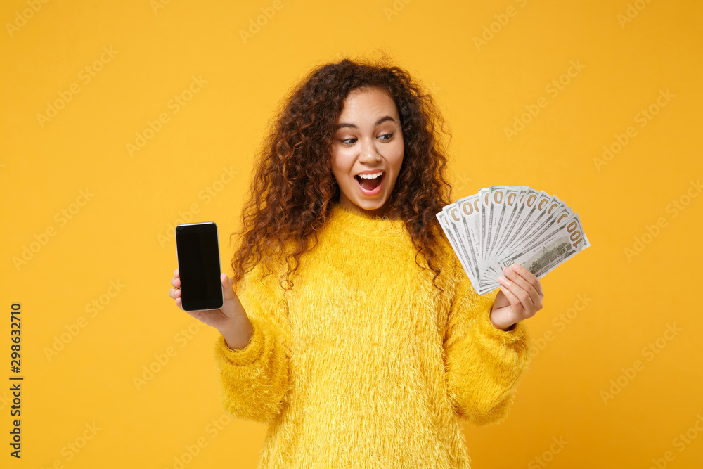 Excited young african american girl posing isolated on yellow background. People lifestyle concept. Mock up copy space. Hold mobile phone with blank empty screen fan of cash money in dollar banknotes. Excited young african american girl posing isolated on yellow background. People lifestyle concept. Mock up copy space. Hold mobile phone with blank empty screen fan of cash money in dollar banknotes.