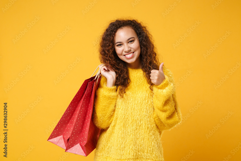 Pretty african american girl in fur sweater posing isolated on yellow orange background. People lifestyle concept. Mock up copy space. Hold package bag with purchases after shopping, showing thumb up. Pretty african american girl in fur sweater posing isolated on yellow orange background. People lifestyle concept. Mock up copy space. Hold package bag with purchases after shopping, showing thumb up.