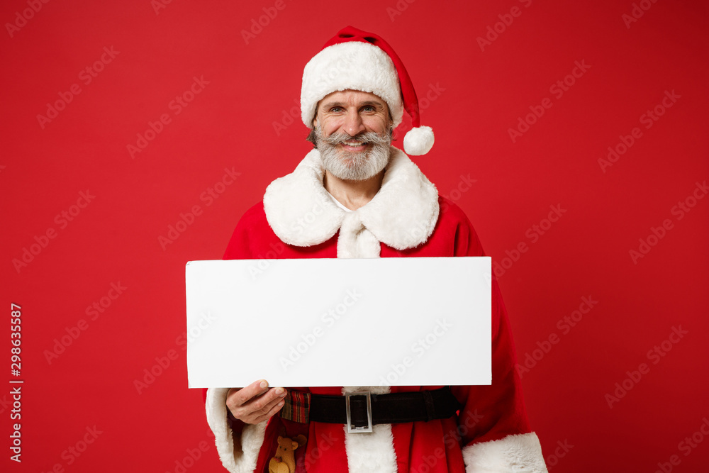 Smiling elderly gray-haired mustache bearded Santa man in Christmas hat posing isolated on red background. New Year 2020 celebration holiday concept. Mock up copy space. Hold white blank sign board. Smiling elderly gray-haired mustache bearded Santa man in Christmas hat posing isolated on red background. New Year 2020 celebration holiday concept. Mock up copy space. Hold white blank sign board.