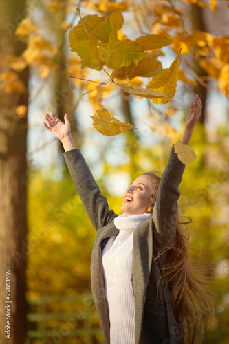 Autumn mood. Happy cheerful girl with outstretched arms throws up yellow leaves. In the autumn park. Attractive smiling young woman with light brown hair in a coat. Vertical portrait. Fall season.