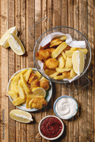 Classic british fast food fish and chips served in frying basket with lemons, red and white sauce, salt in ceramic bowls over wooden plank background. Flat lay, space